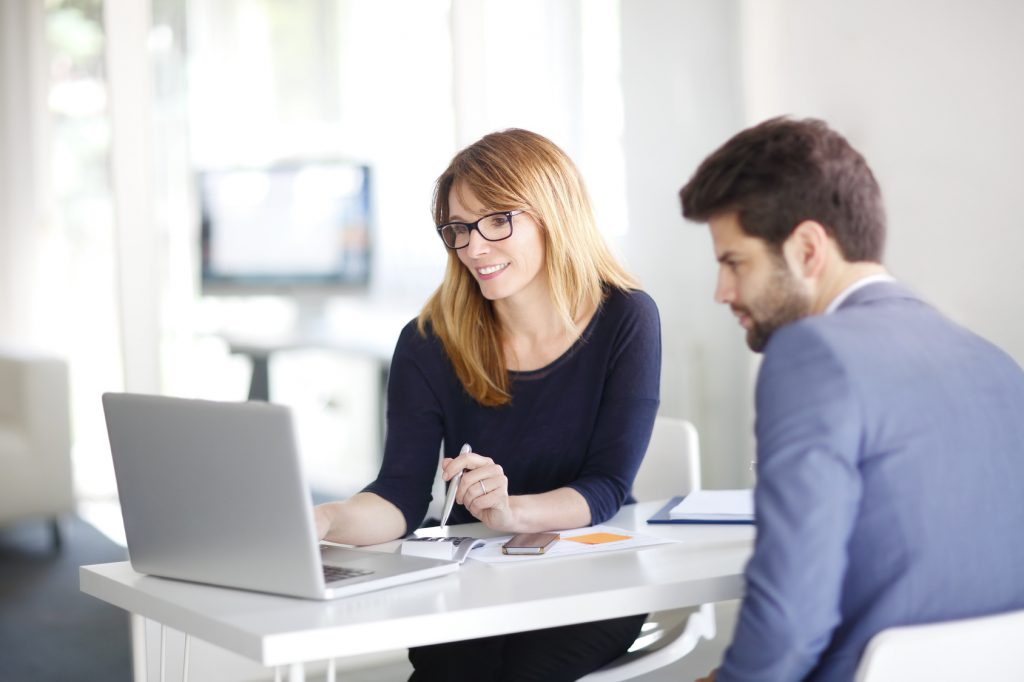 Portrait of investment advisor businesswoman sitting at office in front of computer and consulting with young professional man.